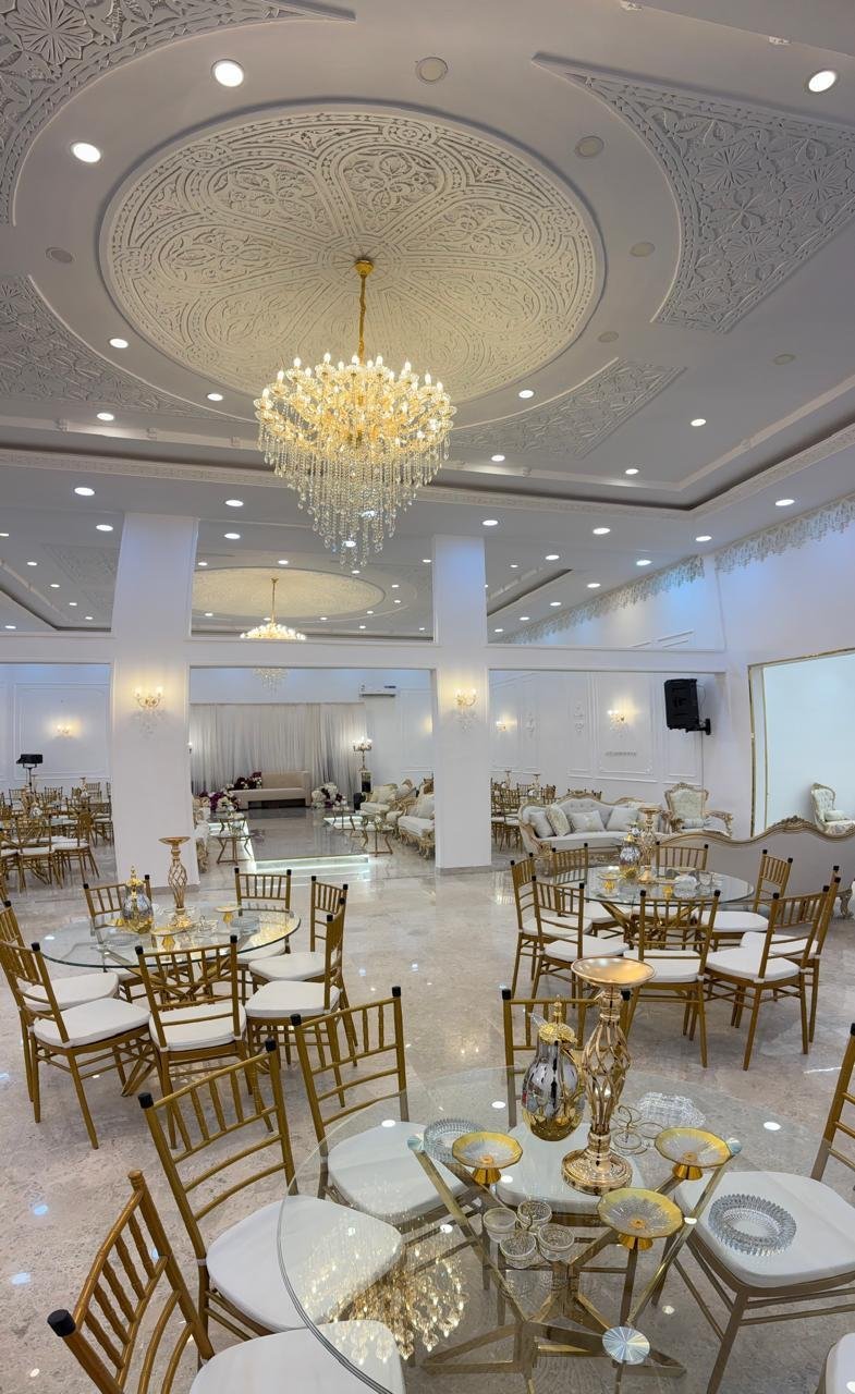 Elegant event venue with ornate ceiling, crystal chandelier, round tables with gold chiavari chairs, and white columns